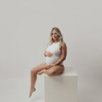 A pregnant woman in a white bodysuit sits gracefully on a white cube against a light background, cradling her belly, captured by a skilled maternity photographer in Phoenix