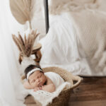 An Arizona newborn photographer captures a peaceful newborn baby resting in a cozy woven basket surrounded by soft, white fabrics and delicate dried floral accents, embodying a sense of serenity and innocence