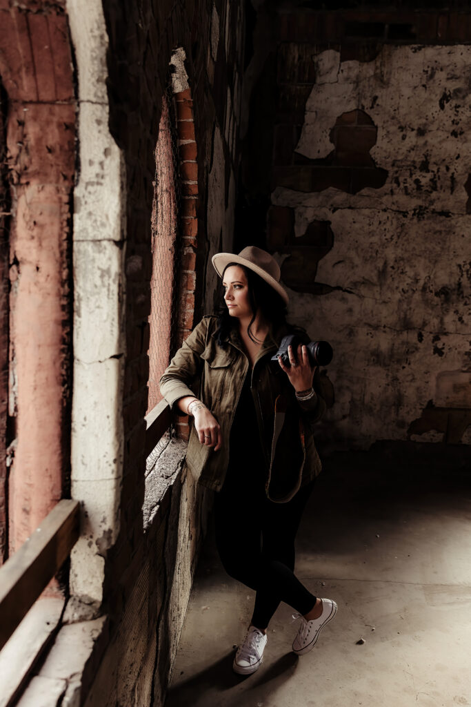 A photographer in a stylish hat and coat holds a camera while thoughtfully gazing out of a rustic window in Arizona, in a room with weathered walls.