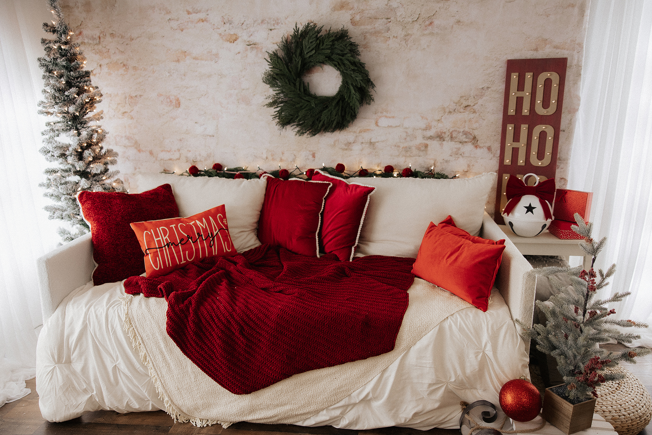 A cozy white sofa with red pillows and a red blanket sits in a festive room decorated for Christmas, featuring a wreath, "Ho Ho Ho" sign, string lights, and small decorated trees.