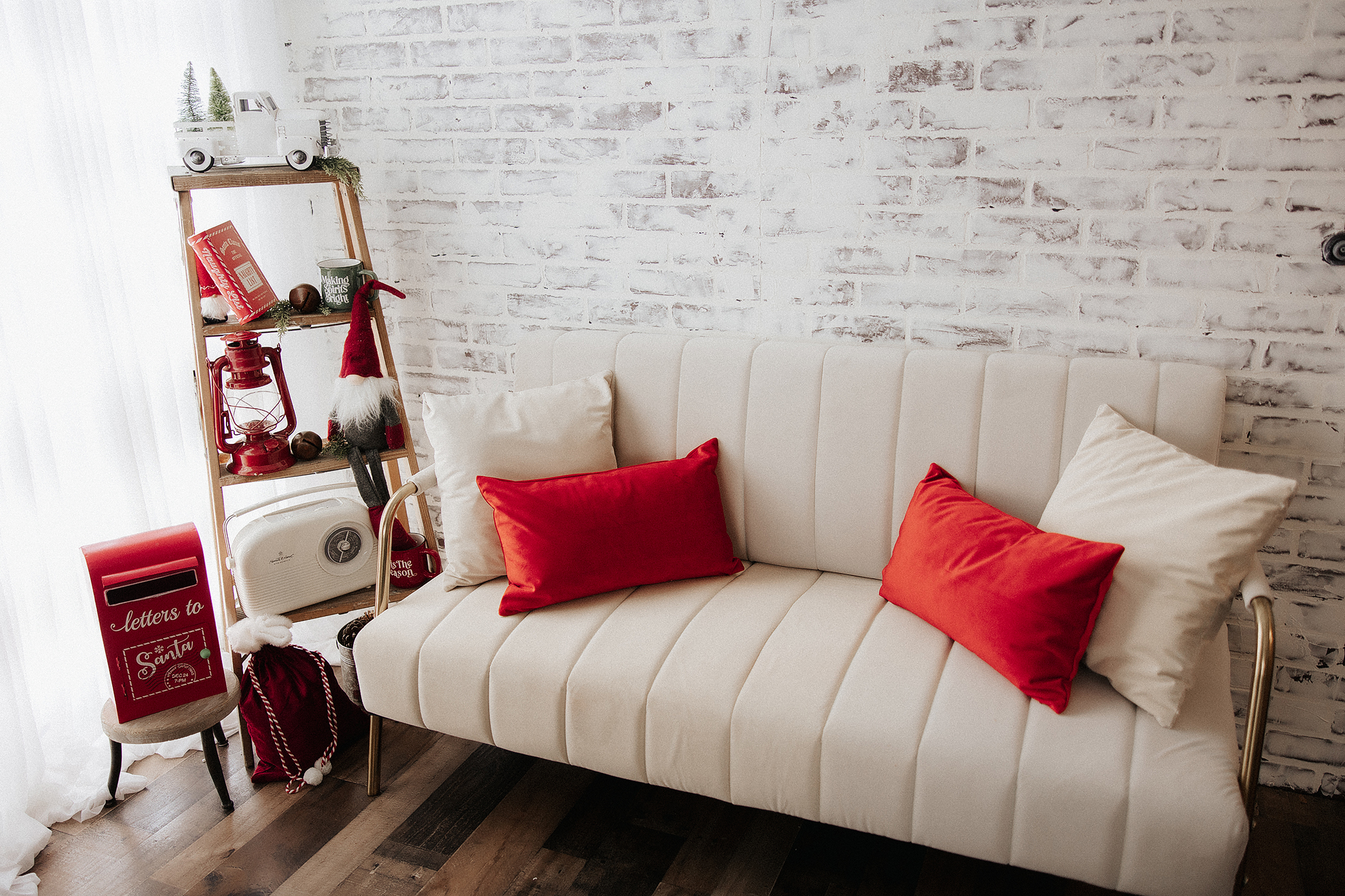 A cozy white sofa with red and cream pillows is set against a white brick wall. Nearby, a shelf holds holiday decorations including a gnome, books, a lantern, and a red mailbox labeled "Letters to Santa.