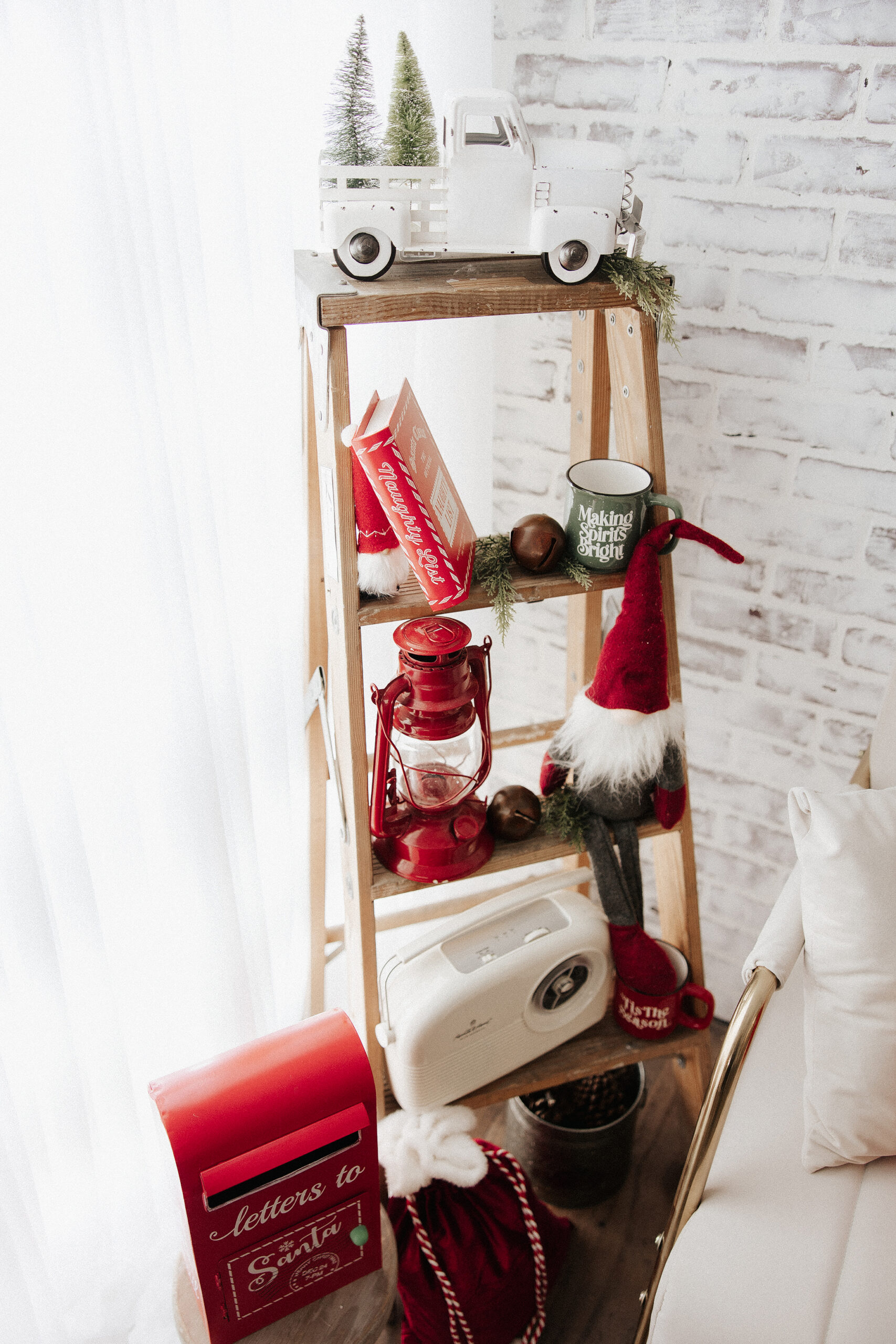 A wooden ladder decorated for Christmas with a white toy truck, mini pine trees, red books, a red lantern, a festive mug, a gnome doll, a vintage radio, a red mailbox, and a red bag on the floor.