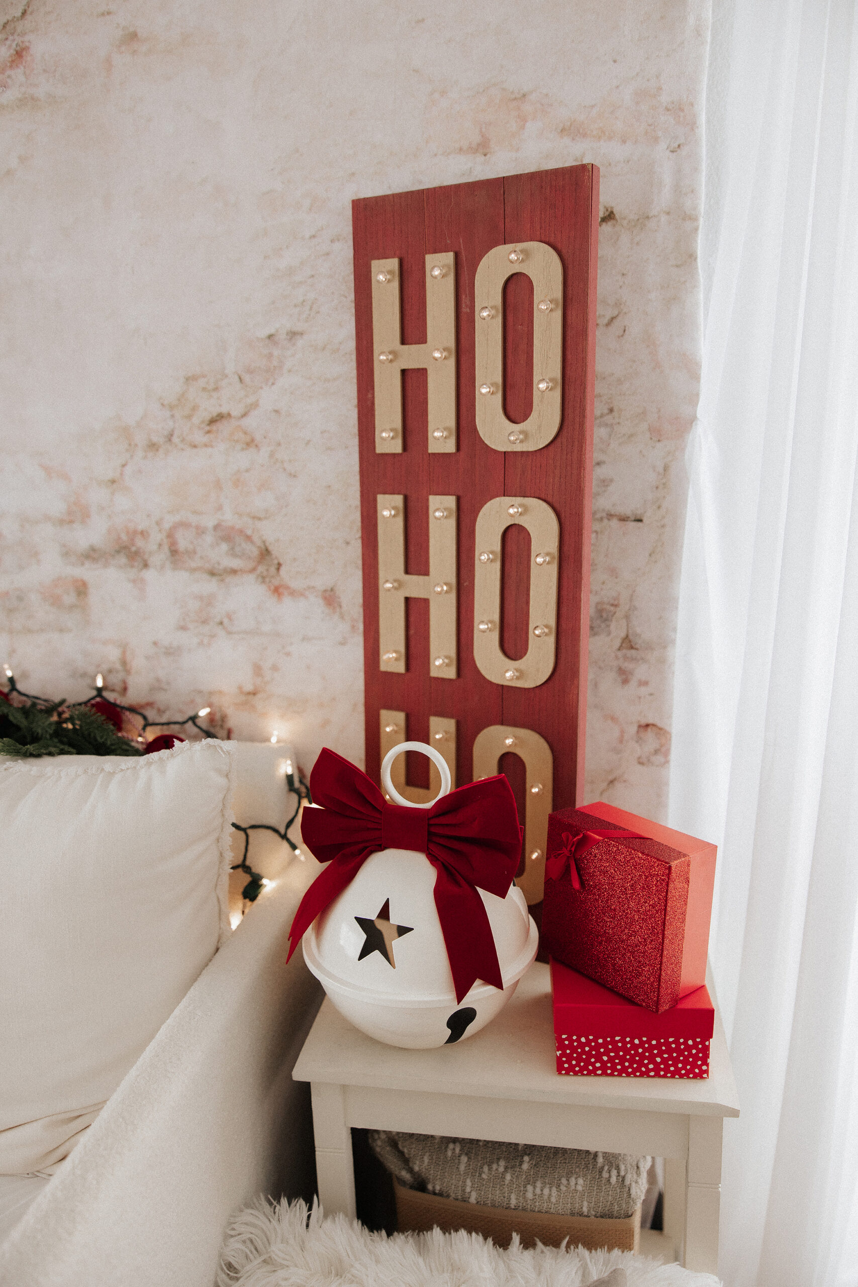 A festive scene with a red “HO HO HO” sign, a large white jingle bell with a red bow, and two red gift boxes on a small table next to a cozy white couch, against a light brick wall.