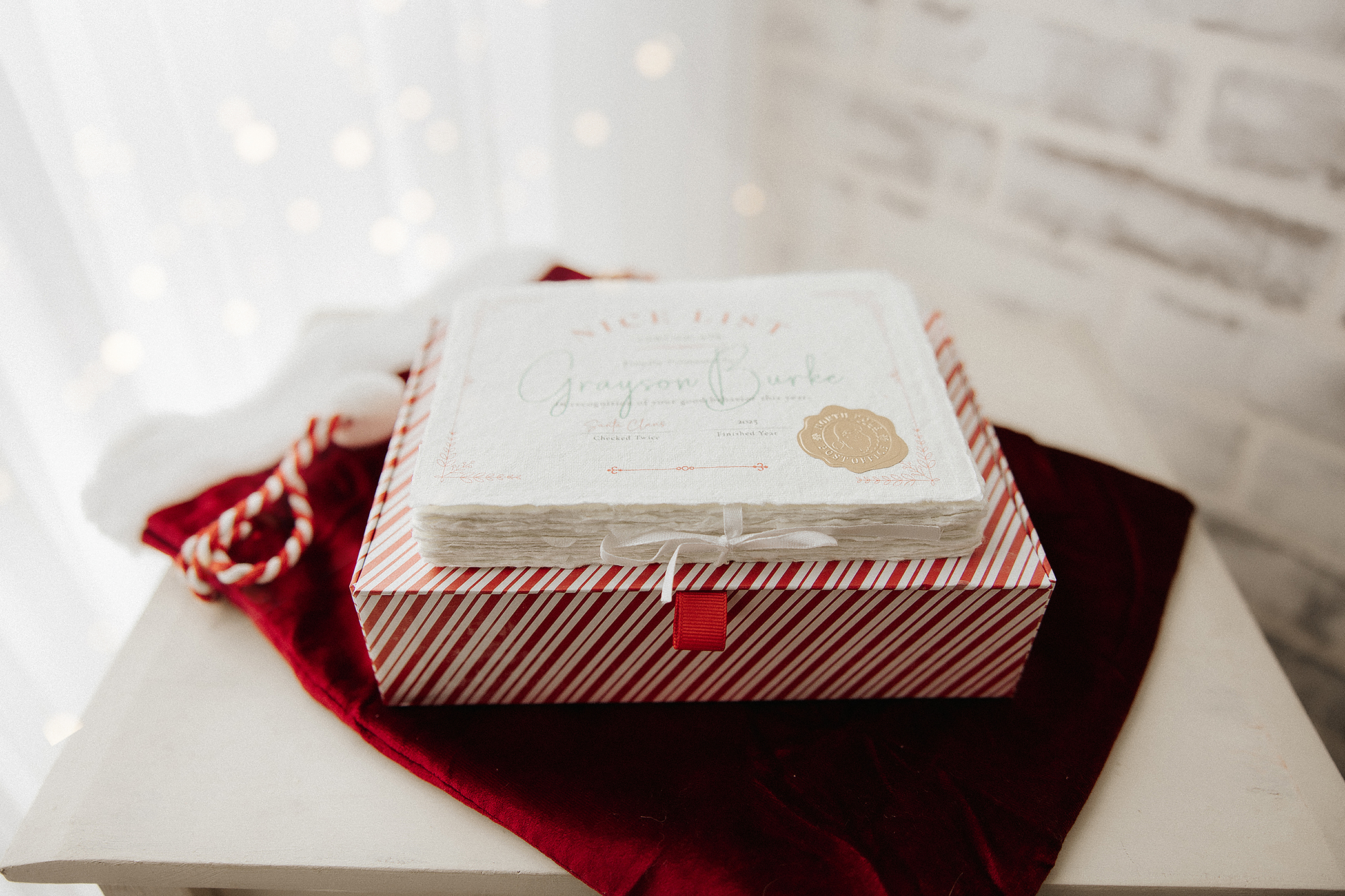 A stack of personalized “Nice List” certificates sits on a red-and-white striped box with a red ribbon, placed on a red velvet cloth and white surface, with candy canes and a white brick wall in the background.