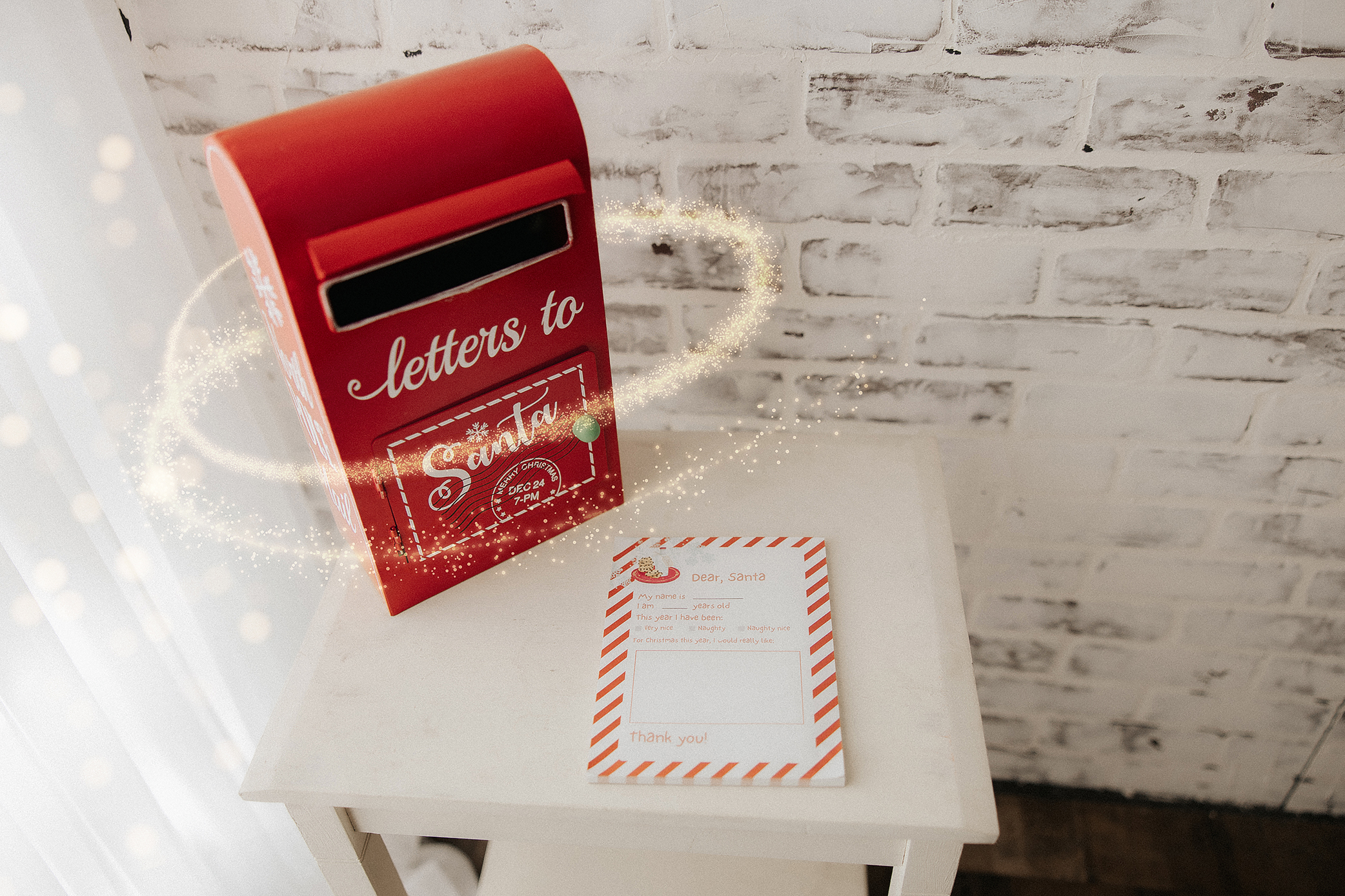 A red mailbox labeled "letters to Santa" sits on a white table next to a festive letter addressed to Santa, with magical sparkles swirling around the mailbox. A white brick wall is in the background.