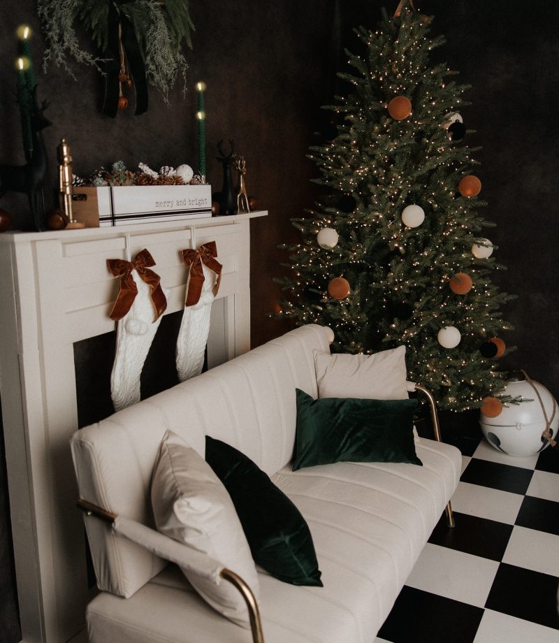 A cozy living room with a white couch, green pillows, and a checkered black-and-white floor. A decorated Christmas tree with white and brown ornaments stands near a white fireplace with stockings and holiday decor.